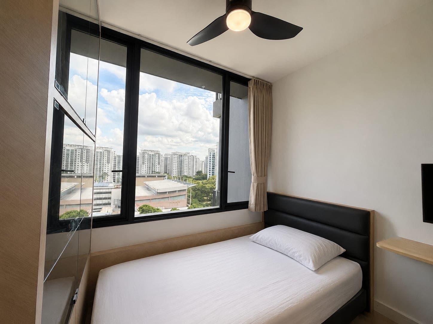 A bedroom with a corner window looking over rooftops and blocks, a pale curtain hanging at the right of the glazing