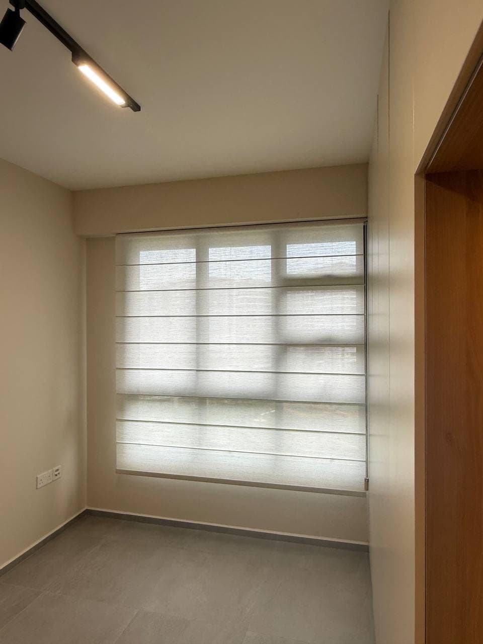 A window fitted with a banded fabric blind beside a timber door frame, track lighting overhead