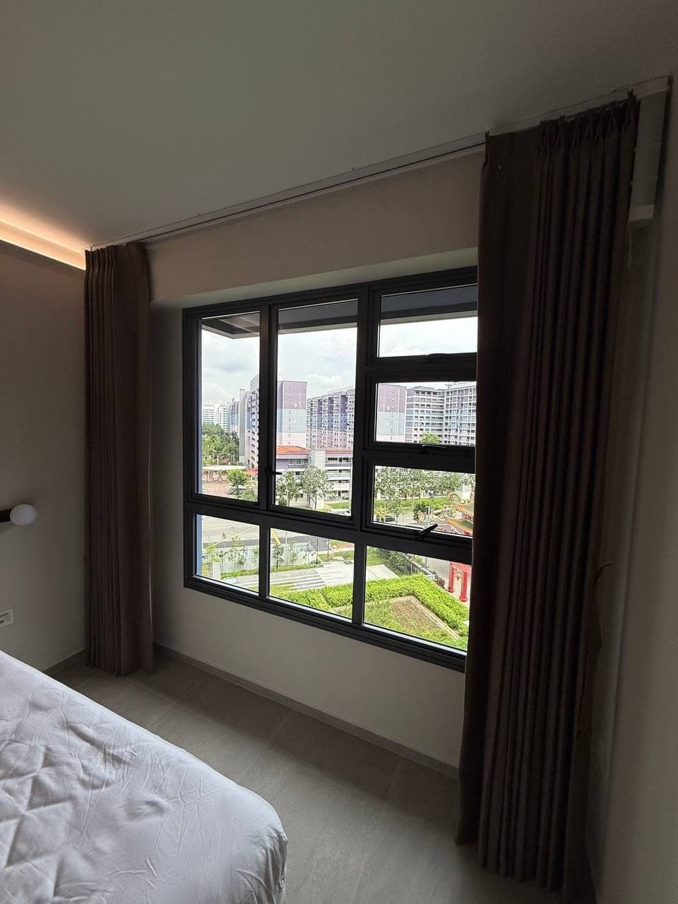 A bedroom with brown curtains at the sides of a window looking onto low blocks and greenery