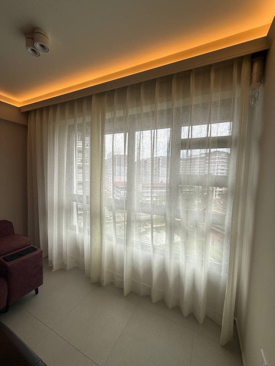 A living area with a cream sheer curtain across the window under a cove-lit ceiling, sofa at the left
