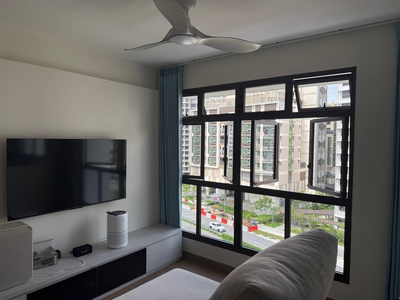 A living room with a wall-mounted television, ceiling fan and windows screened by pale curtains