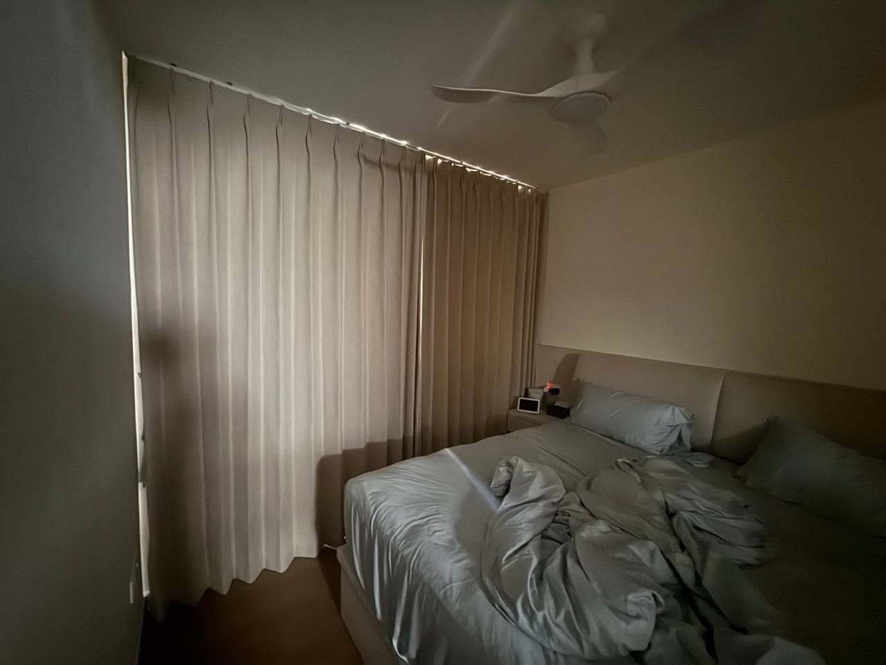 A dimly lit bedroom with a cream curtain drawn across the window and a made bed in the foreground